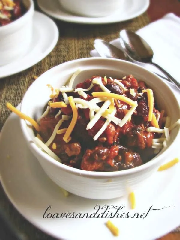 A close up view of a white bowl of party chili with two spoons and shredded cheese on top