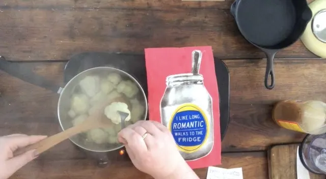 Cauliflower cooking in water in a pan, hand stirring, pink towel in background