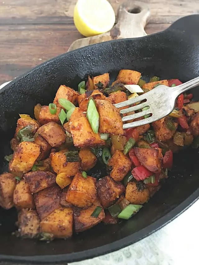 A fork picking up a piece of the sweet potato hash from an oval cast iron pan with wood in the background