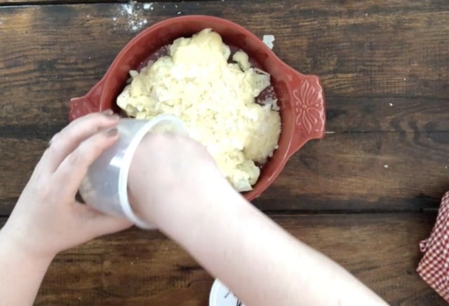 hand sprinkling parmesan over the cooked cauliflower with cheese sauce that is in a red baking dish