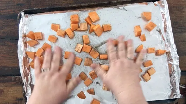 Hands spreading cubed sweet potatoes out on an oiled half sheet pan covered with foil