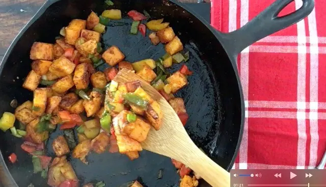 A wooden spoon scooping out some of the sweet potato hash browns from a skillet with red and white napkin in the background
