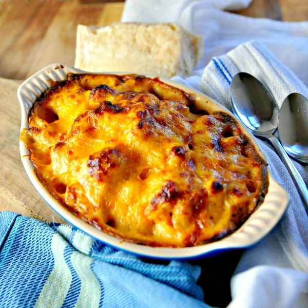 Oval baking dish filled with browned cauliflower cheese, sitting on blue napkins with parmesan block in background and two spoons