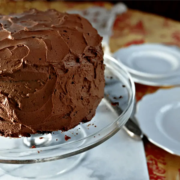 Homemade chocolate cake on a glass cake plate with white plates in the background