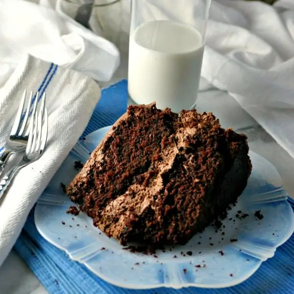 a piece of chocolate cake sitting on a see through white antique dish with a glass of milk behind and two forks