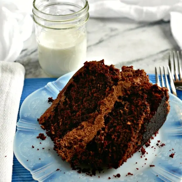 a close up photo of homemade chocolate cake on a white plate with crumbs, two forks and a half pint jar of milk