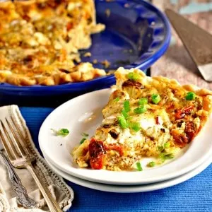 A slice fo Roasted Tomato and Garlic Quiche on a white plate with a blue placemat and two forks
