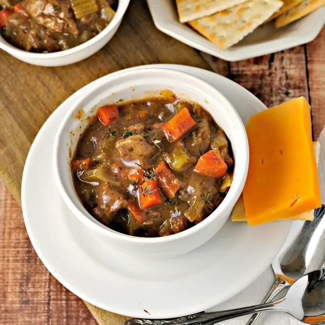 A small white bowl of beef stew sitting on a white saucer with a piece of yellow cheese beside it