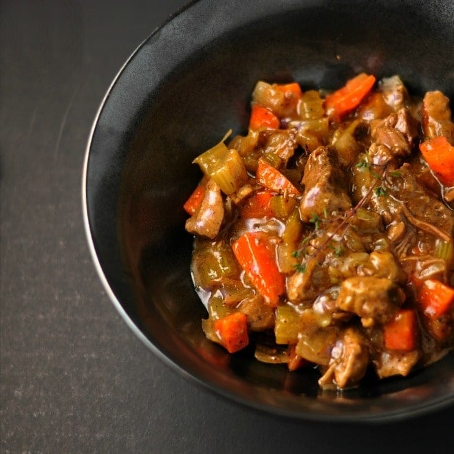 A black bowl sitting on a dark gray background. The bowl is full of classic beef stew that is shiny and full of orange carrots