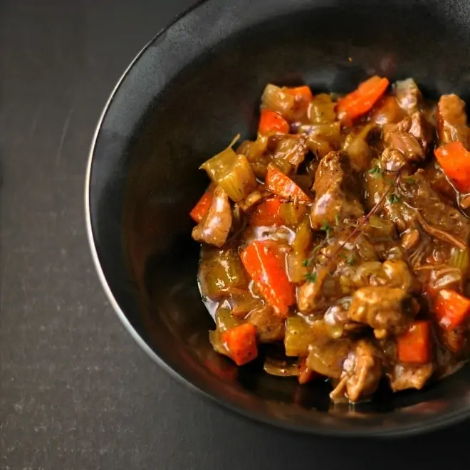 A black bowl sitting on a dark gray background. The bowl is full of classic beef stew that is shiny and full of orange carrots