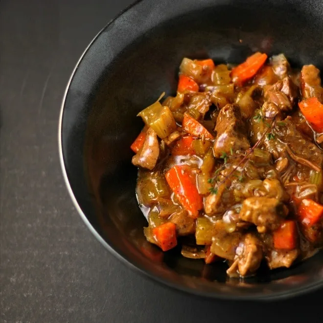 A black bowl sitting on a dark gray background. The bowl is full of classic beef stew that is shiny and full of orange carrots