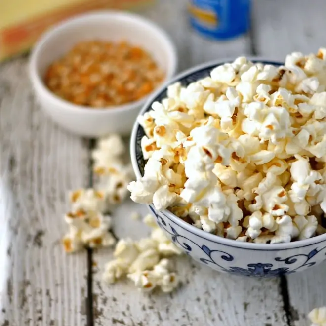 A bowl of popcorn on a white table with a small bowl of popcorn kernels in the background