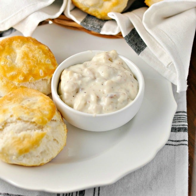 A photo of a bowl of gravy sitting on a small plate with two biscuits