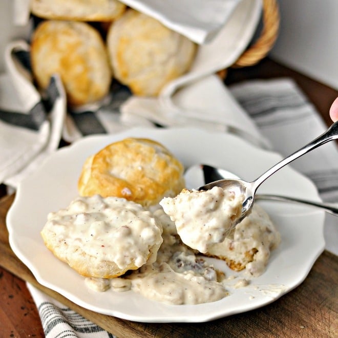 A plate of gravy on two biscuits with someone holding a spoonful of the gravy up to the camera