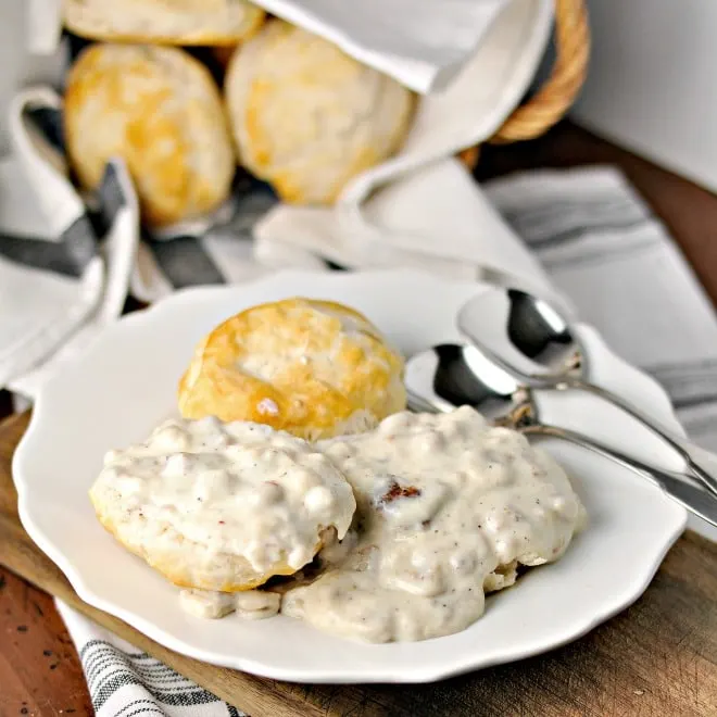 A white plate with an open face biscuit with gravy on top of it and there are two spoons on the plate