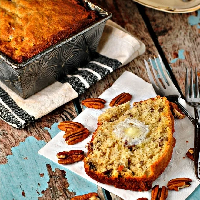 Slice of banana bread with melted pat of butter, pecans, fork and loaf surrounding 