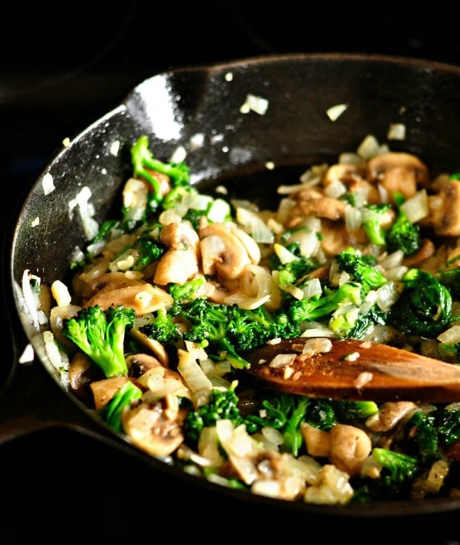 Broccoli, mushrooms, onions and garlic being sautéed in a cast iron skillet with a wooden spoon