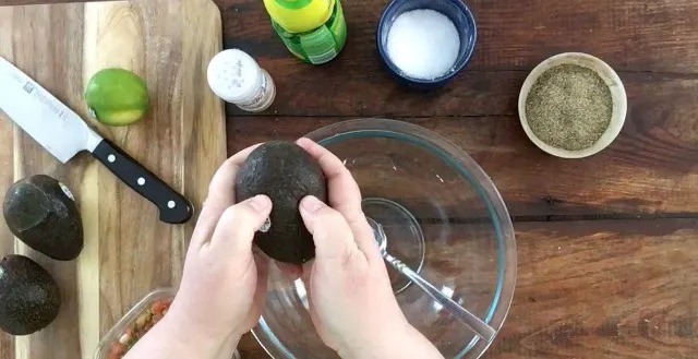 two hands squeezing an avocado over a glass bowl