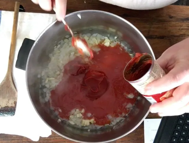 A photo of a hand adding tomato past to a large stock pot with onions and garlic using a spoon