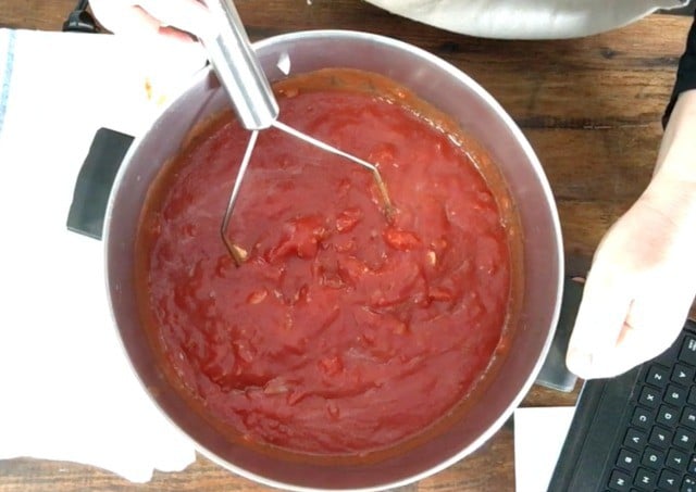 A potato masher in a large stock pot with the tomato sauce