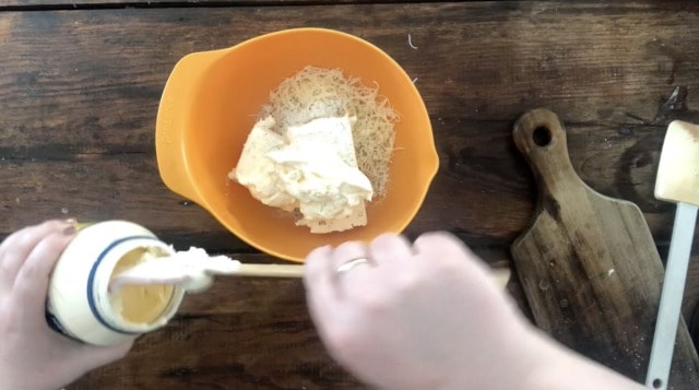 hands adding mayonnaise to an orange bowl of ingredients.