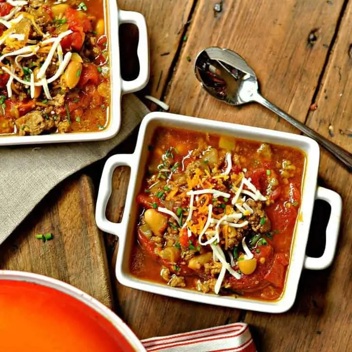 two square bowls filled with colorful chili sprinkled with cheese. Brown napkin and striped towel, spoon and orange pot lid in background