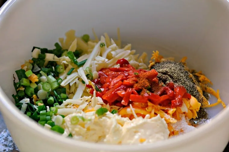 White bowl with all of the ingredients in it showing green onions, white mayo, red pimentos and yellow cheese.