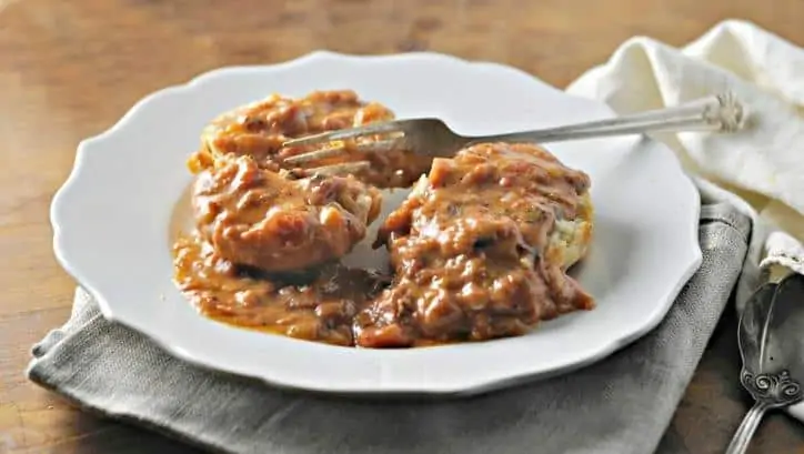 a fork digging into a biscuit covered with tomato gravy on a white plate with a brown linen napkin