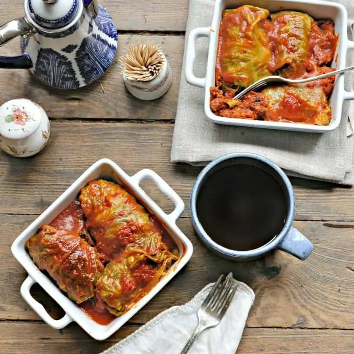 two square white bowls with handles holding three cabbage rolls each, coffee cup in back ground