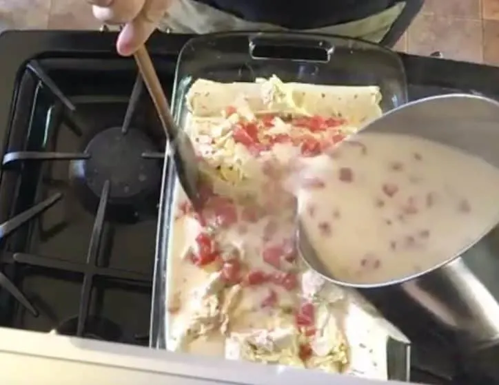 cheese sauce being poured over the enchiladas in a baking dish