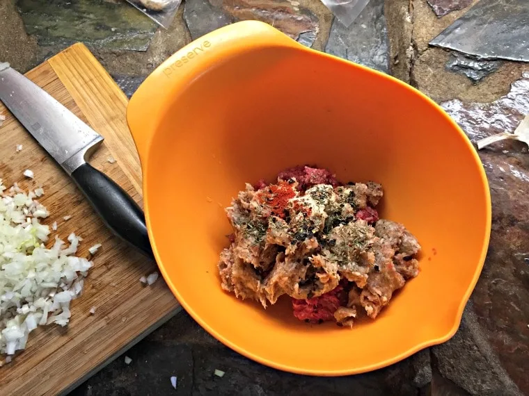 An orange mixing bowl with meat mixture inside and chopped onion in background