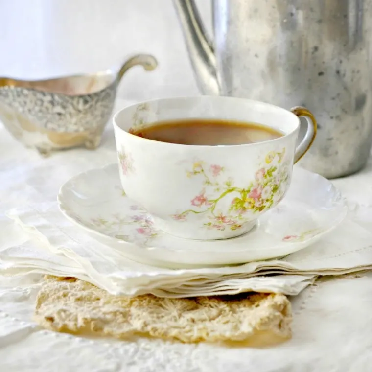 A cup of tea in a bone china cup with saucer, and linen cloth napkins with silver tea pot and sugar bowl in back ground