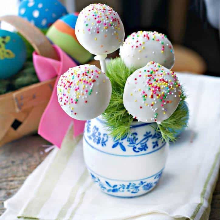 white jar with blue flowers holding 4 cake pops, eggs in background. white and green napkin underneath