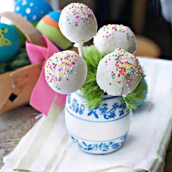 white jar with blue flowers holding 4 cake pops, eggs in background. white and green napkin underneath