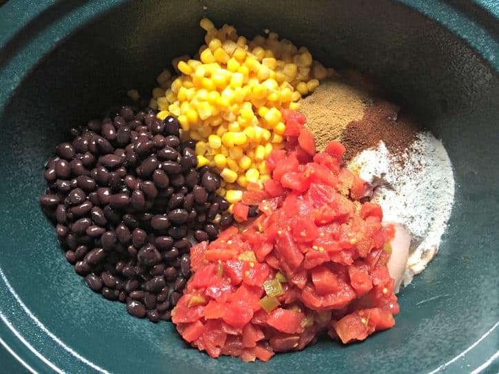 Crock pot bowl with tomatoes, black beans, corn and seasonings on top of the breasts.