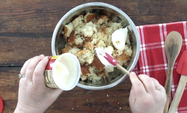 Hand scraping cream cheese icing from can into bowl of crumbled cake.