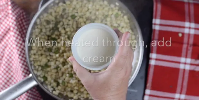 A small white bowl of cream being held over the pan of corn and jalapenos