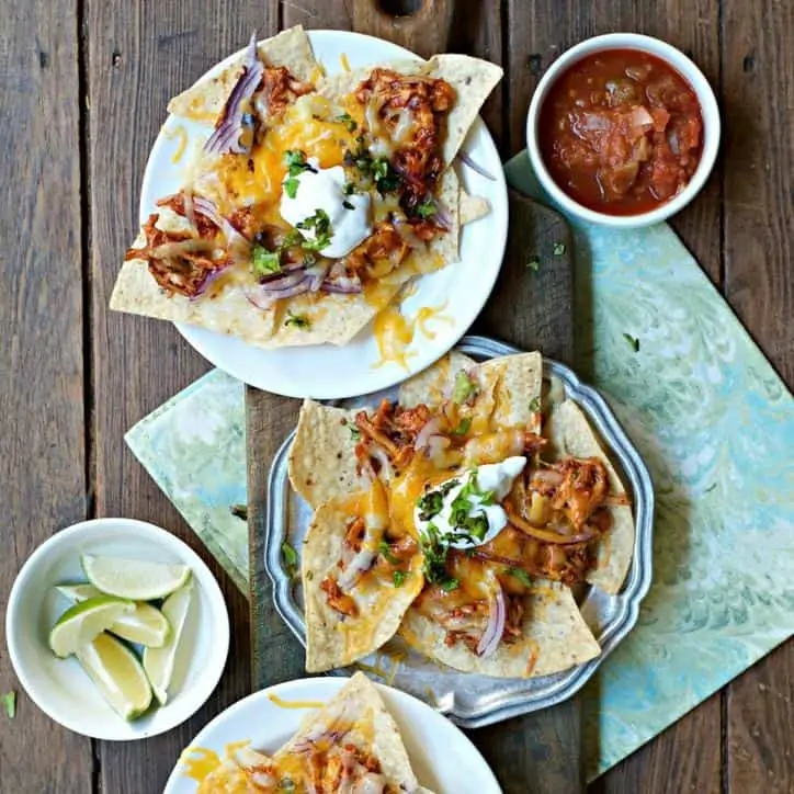 Three plates of nachos topped with sour cream sitting on green swirled napkin with lime and salsa to side