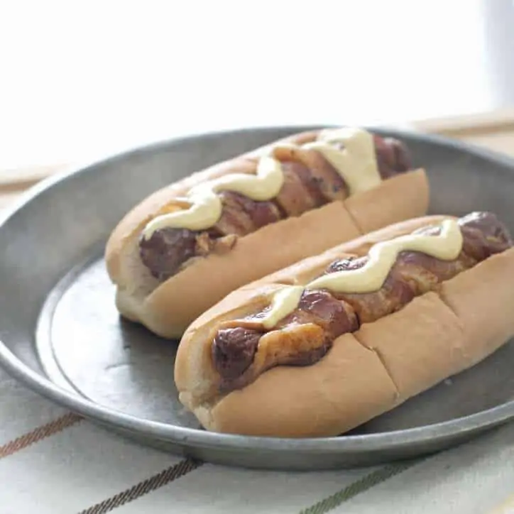 Two bacon wrapped hot dogs on a gray plate sitting on a striped towel with sunlight through window