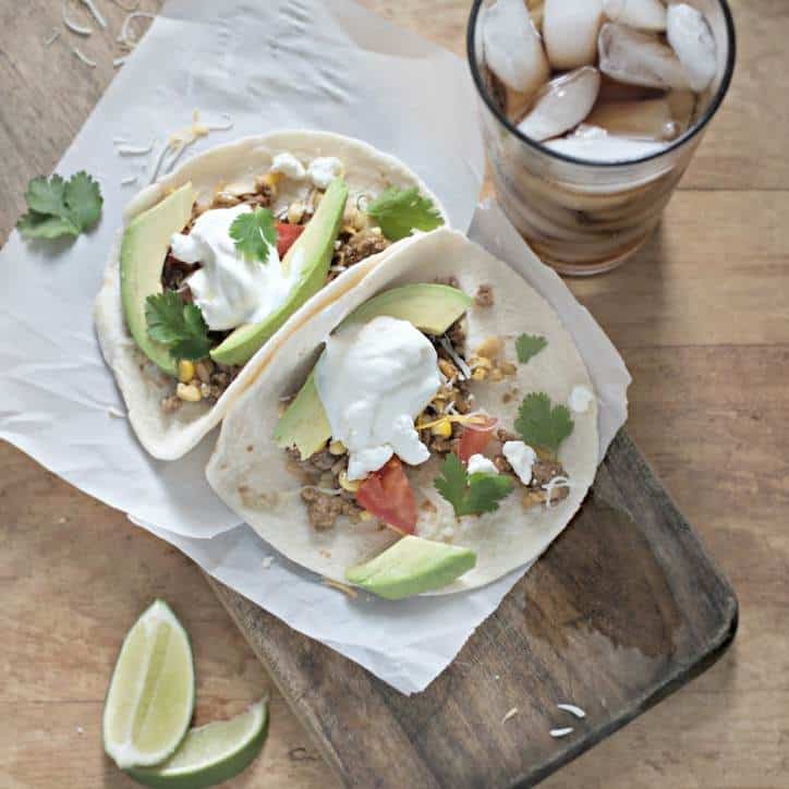 Two soft tacos filled with ground beef, corn, avacado, sour cream and cilantro on wax paper on a cutting board with coke and lime to the side