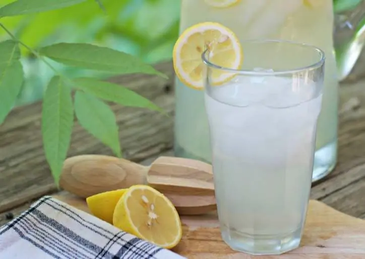 A close up photo of the lemonade glass with lemon on the rim and pitcher in the background