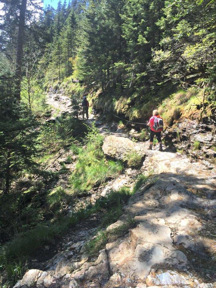 A photo of two hikers on a small path with large slopes and rocks to the left.