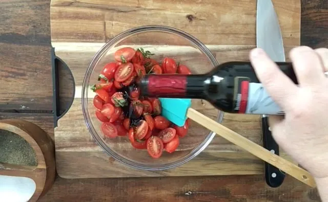 A bottle of balsamic vinegar being poured into the salad, blue spatula mixing in clear bowl on cutting board