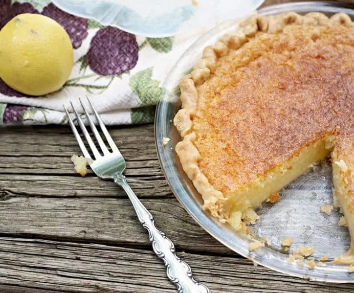 This is a photo of a Old Fashioned Lemon Chess Pie with a slice cut out a fork a lemon and a kitchen towel