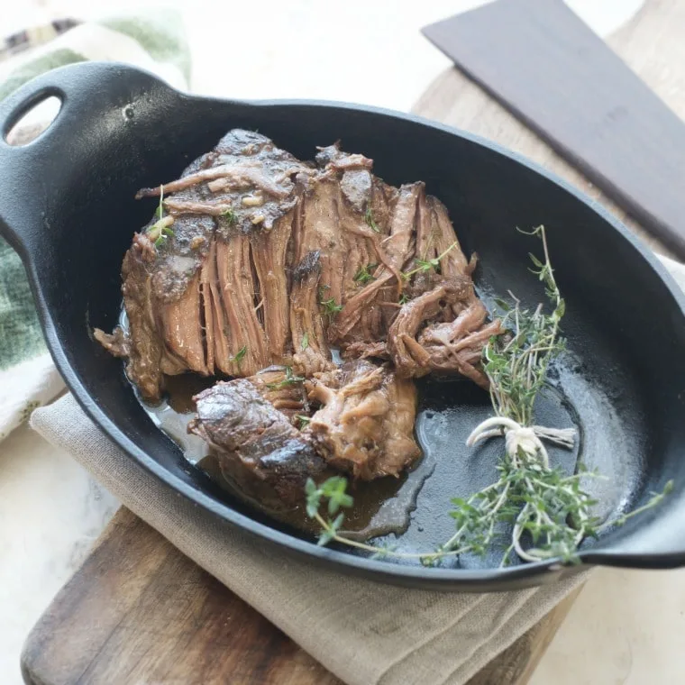 An overhead and to the side view of the crock pot roast recipe in an oval cast iron pan with thyme bundle and cutting board in the back ground