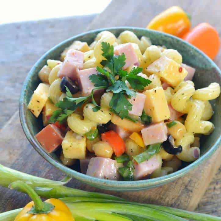 An overhead view of a green bowl of pasta salad sitting on cutting board with onion and peppers. 