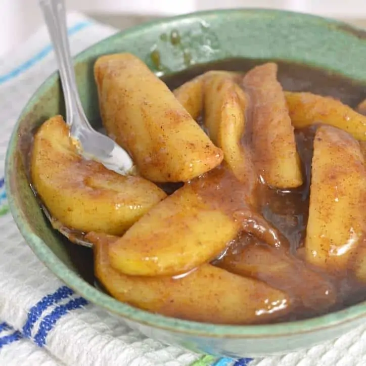 A photo of a green bowl of fried apples with a spoon in the bowl and resting on a white towel with blue stripes