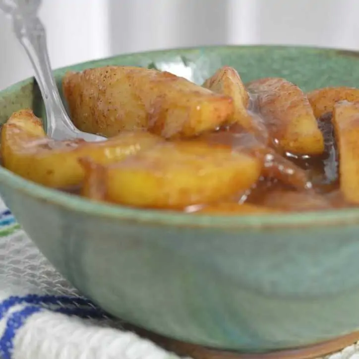 A photo of a green bowl of fried apples and spoon sitting on a white towel with blue stripes from the side THE SECRET TO PERFECT SOUTHERN FRIED APPLES