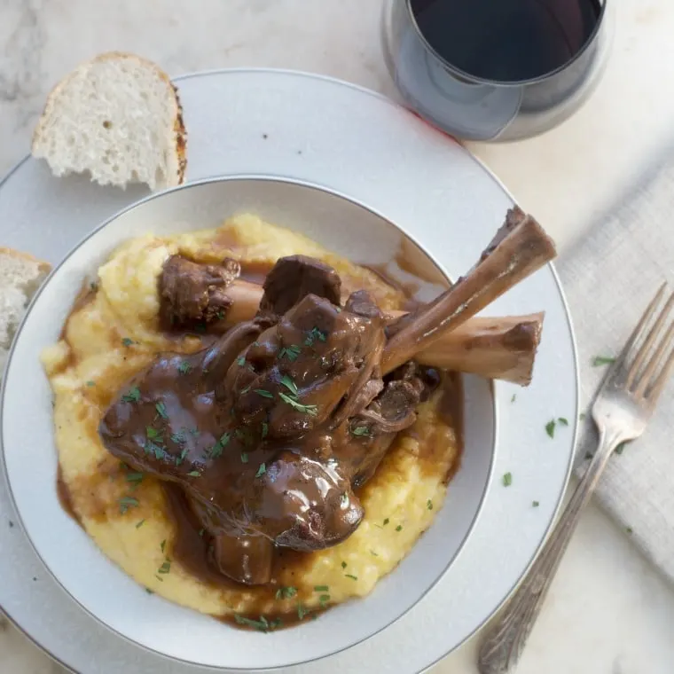 Lamb Shanks over polenta made with gravy in bowl on plate with bread half, fork and wine