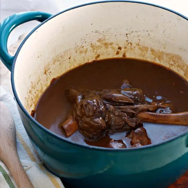 Blue dutch oven with towel in back ground and wooden spoon. Cooked Lamb Shank in the brown gravy